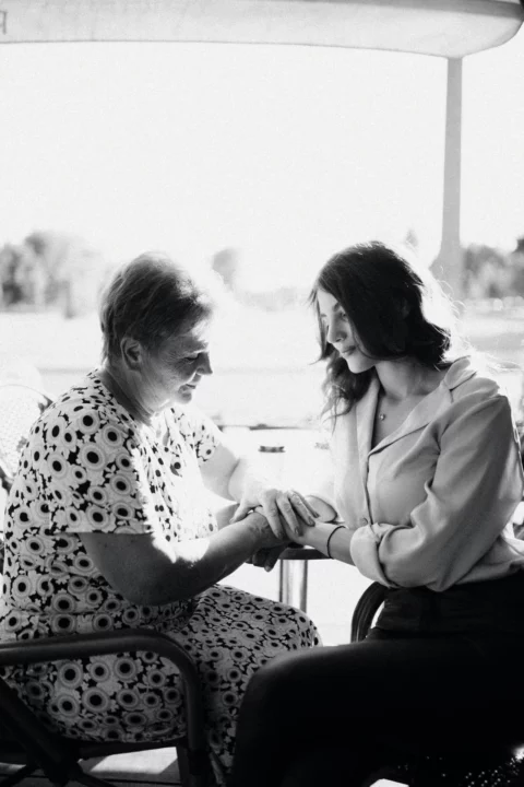 Younger woman hand-in-hand with older woman comforting here by a large window